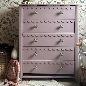 May include: A mauve-colored dresser with five drawers, each featuring a scalloped edge. The drawers have decorative floral knobs. A wooden toy horse sits on top. The dresser is in a room with floral wallpaper.