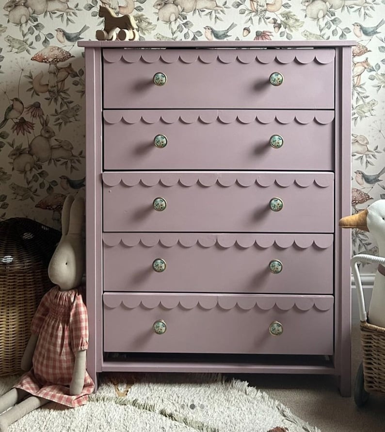May include: A mauve-colored dresser with five drawers, each featuring a scalloped edge. The drawers have decorative floral knobs. A wooden toy horse sits on top. The dresser is in a room with floral wallpaper.