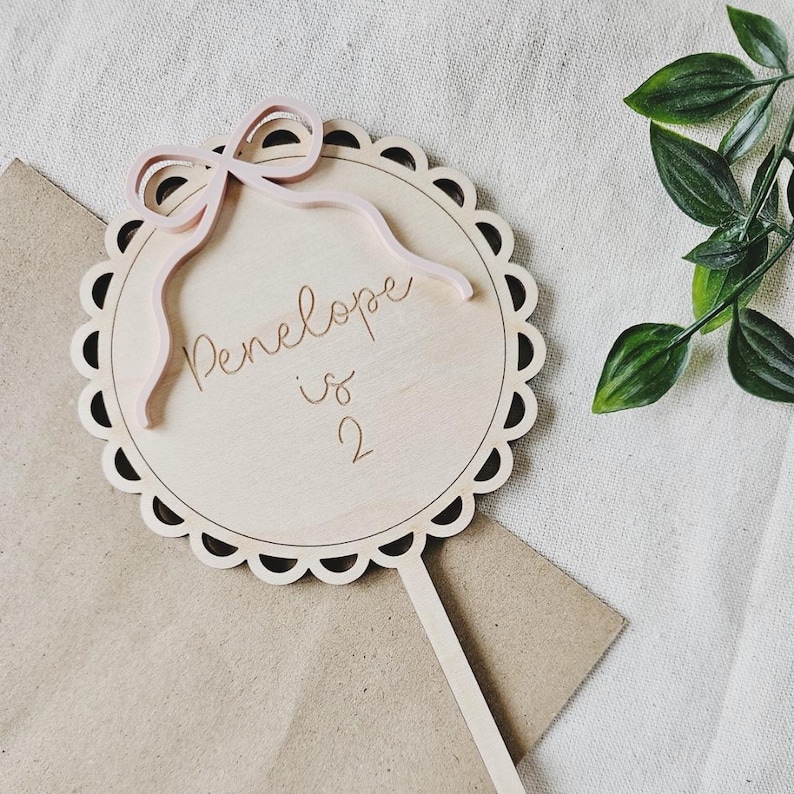 May include: A wooden cake topper with a scalloped edge and a pink bow. The topper says "Penelope is 2".