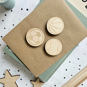 May include: Three wooden sports ball magnets, a soccer ball, a basketball, and a baseball. The magnets are on a brown surface.