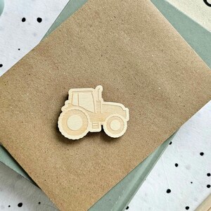 May include: A light-coloured wooden cutout in the shape of a tractor featuring a detailed outline of the cab, engine, and large wheels. The tractor cutout is positioned on a textured brown cardstock surface with a visible grain and smooth finish.