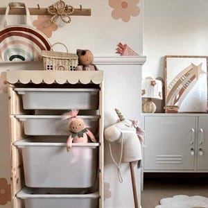 May include: A wooden storage unit with four white plastic drawers. The unit has a scalloped edge and is decorated with pastel pink and brown flower decals. A pink doll, a stuffed unicorn, and a wicker basket are on the shelves. The text "Reach for the Stars" is visible on a framed rainbow print.