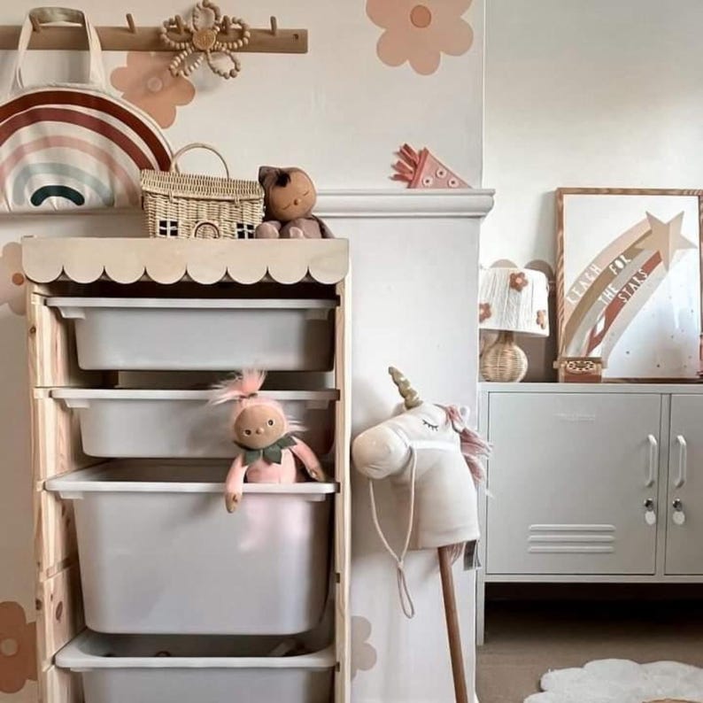 May include: A wooden storage unit with four white plastic drawers. The unit has a scalloped edge and is decorated with pastel pink and brown flower decals. A pink doll, a stuffed unicorn, and a wicker basket are on the shelves. The text "Reach for the Stars" is visible on a framed rainbow print.