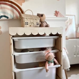 May include: A white wooden toy storage unit with scalloped edges on the top shelf. The unit has four white plastic drawers. A pink doll with green leaves around its neck is peeking out of the top drawer.
