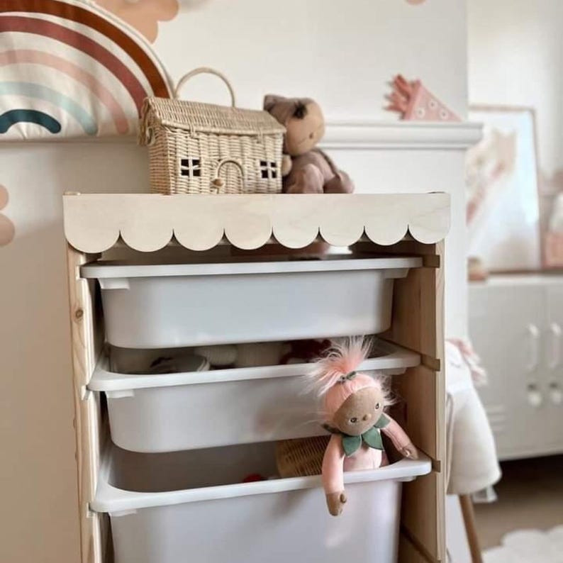 May include: A white wooden toy storage unit with scalloped edges on the top shelf. The unit has four white plastic drawers. A pink doll with green leaves around its neck is peeking out of the top drawer.