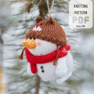 May include: A knitted snowman ornament with a brown hat, red scarf, and orange beak hangs from a pine branch. The snowman has black button eyes and a smiling face. The text "KNITTING PATTERN PDF Only ENG" is in the upper right corner.