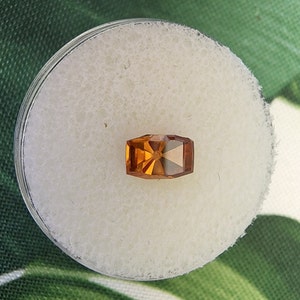 May include: A rectangular, orange gemstone with a faceted cut. The gemstone is set in a clear, round container.