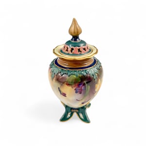 Könnte beinhalten: Eine dekorative Keramikurne mit Deckel, mit einem Blumendesign in Lila-, Gelb- und Grüntönen. Die Urne hat eine goldfarbene Spitze und einen grünen Sockel. Der Deckel hat ein rosa-grünes Design.