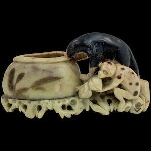 Può includere: Una scultura in pietra scolpita raffigurante un orso nero e una scimmia maculata che interagiscono con un piccolo vaso rotondo. Il vaso è verde chiaro con disegni marroni a forma di foglia. La scultura poggia su una base decorativa con dettagli traforati. Lo sfondo è nero.