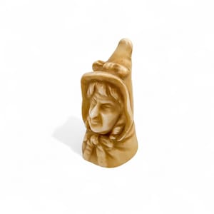 May include: A tan-colored ceramic figurine depicting a person wearing a hat and a hooded cloak. The figurine has detailed facial features and is set against a white background. The figure's face is turned to the side.