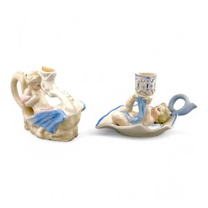 May include: Two porcelain candle holders featuring cherubs. One cherub is sitting on a swan, holding a candle. The other cherub is lying on a leaf-shaped base, holding a candle in a blue and white cup.