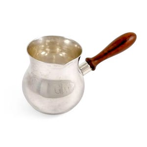 May include: A silver creamer with a wooden handle. The creamer has a rounded body and a small spout. The handle is dark brown and has a smooth finish.