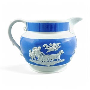 Puede incluir: Jarra de cerámica azul y blanca con asa y boquilla. La jarra presenta un cuerpo azul con detalles en relieve blancos de querubines y un carro. Una banda blanca rodea la parte superior.