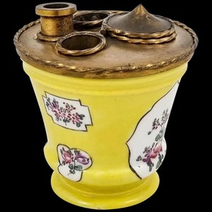 Puede incluir: Un tintero de porcelana amarillo con una tapa de latón y tres anillos de latón. El tintero está decorado con motivos florales en rosa, verde y blanco.