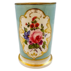 Puede incluir: Un jarrón de cerámica azul claro con un diseño floral. El jarrón presenta un panel central con una rosa rosa, flores rojas y moradas y flores azules. Los adornos dorados acentúan la parte superior e inferior.