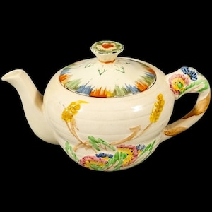 Puede incluir: Una tetera blanca con un diseño floral en tonos de amarillo, verde, azul y rosa. La tetera tiene una tapa con un diseño floral y un pico en el lateral.