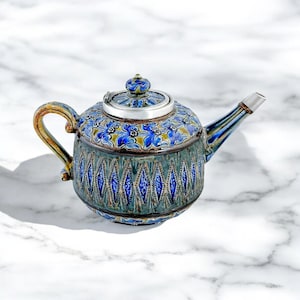 Puede incluir: Una tetera de cerámica azul y verde con una tapa de plata y un patrón floral. La tetera tiene un diseño en forma de diamante alrededor de la parte inferior.