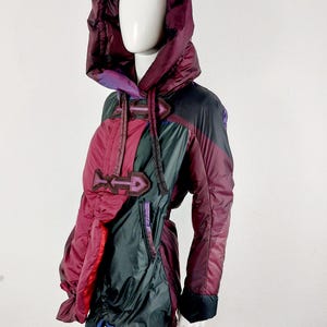 Op de afbeelding: Een veelkleurige jas met capuchon, getoond op een mannequin. De jas heeft tinten bordeaux, zwart en groen. De jas heeft een gewatteerd ontwerp, een grote capuchon en decoratieve details. Het heeft een uniek, asymmetrisch ontwerp met een mix van texturen en kleuren.