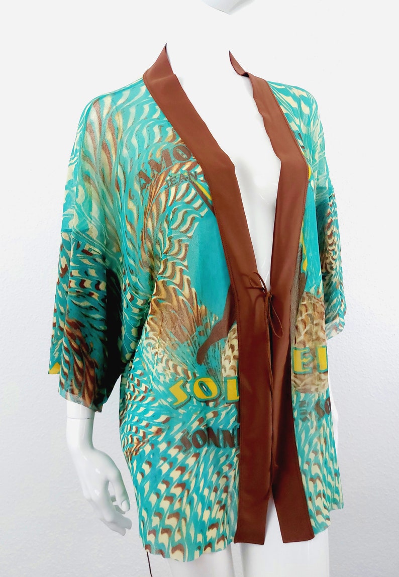 May include: A turquoise and brown sheer kimono with a brown satin trim and tie closure. The kimono has a colorful print of feathers and the words "Soleil" and "Amore" on the front.