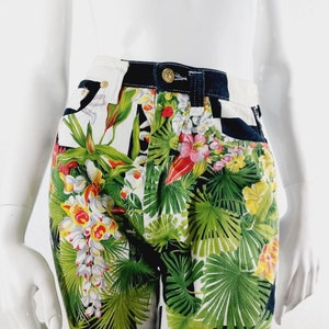 May include: High-waisted denim shorts with a tropical floral print featuring green leaves, white and yellow flowers, and red accents. The shorts have a black waistband and a checkered pattern on the back.