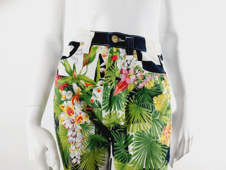 May include: High-waisted denim shorts with a tropical floral print featuring green leaves, white and yellow flowers, and red accents. The shorts have a black waistband and a checkered pattern on the back.