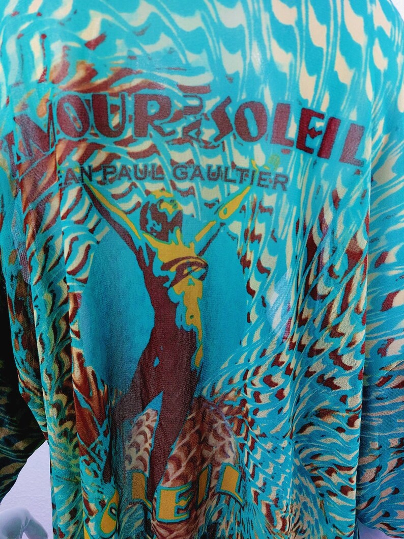 May include: A turquoise blue sheer top with a brown and yellow graphic print of a woman with the text "Amour Soleil Jean Paul Gaultier" on the front.
