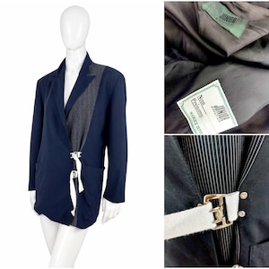 May include: Navy blue blazer with a gray panel and white strap detail. The blazer features a unique design with a contrasting panel on the front and a white strap with a metal buckle closure. The blazer is displayed on a mannequin.