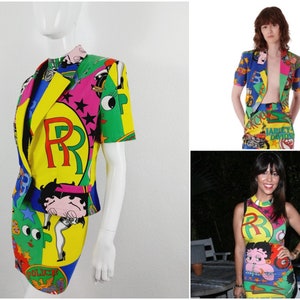 May include: A colourful, short-sleeved jacket and skirt set with a cartoon print featuring a large "R" and a cartoon character. The set is made of a bright yellow, green, blue, pink, and red fabric.
