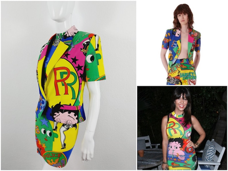 May include: A colourful, short-sleeved jacket and skirt set with a cartoon print featuring a large "R" and a cartoon character. The set is made of a bright yellow, green, blue, pink, and red fabric.