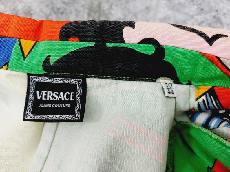 May include: A close-up of a white, green and black patterned fabric with a black label that reads "VERSACE JEANS COUTURE". A smaller white label with black text reads "30 44".