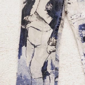 May include: A pair of blue denim jeans with a white background featuring a black and white illustration of a man in a military uniform. The illustration is faded and has a vintage look.