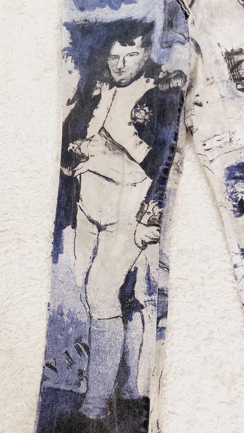 May include: A pair of blue denim jeans with a white background featuring a black and white illustration of a man in a military uniform. The illustration is faded and has a vintage look.