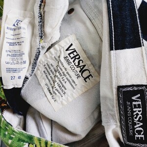 May include: Close-up of a white label with black text that reads "VERSACE JEANS COUTURE". The label is attached to a pair of black and white patterned jeans with a tropical print.