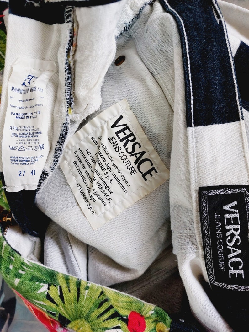 May include: Close-up of a white label with black text that reads "VERSACE JEANS COUTURE". The label is attached to a pair of black and white patterned jeans with a tropical print.