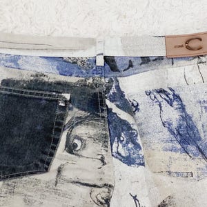 May include: White denim jeans with a blue and white abstract print. The jeans have a dark blue pocket and a brown leather patch with the text "Occult" and a stylized "C" logo.