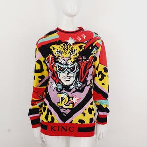 Puede incluir: Un vibrante suéter de manga larga con un diseño de estilo pop art. El suéter presenta un gráfico de una persona con gafas de sol y un tocado de leopardo, con la palabra "KING" en la parte inferior. La paleta de colores incluye rojo, amarillo, negro y rosa.
