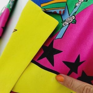 May include: A colourful blazer with a patchwork design featuring stars, flowers, and geometric shapes. The blazer is made of a smooth fabric and has a yellow, pink, blue, green, and red colour scheme.