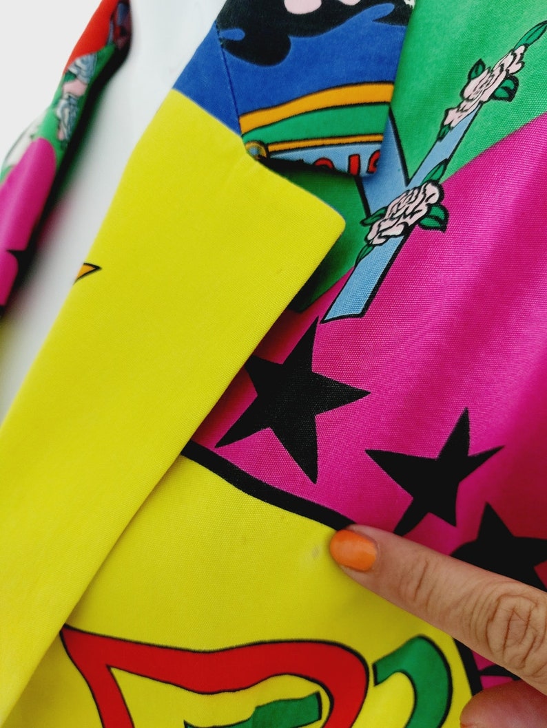 May include: A colourful blazer with a patchwork design featuring stars, flowers, and geometric shapes. The blazer is made of a smooth fabric and has a yellow, pink, blue, green, and red colour scheme.