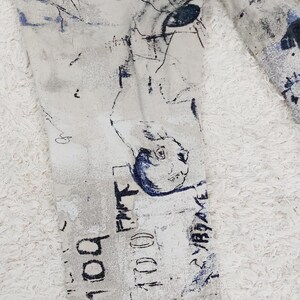 May include: A pair of white denim jeans with a blue and black abstract print. The print features a variety of shapes and lines, including a face with an eye.