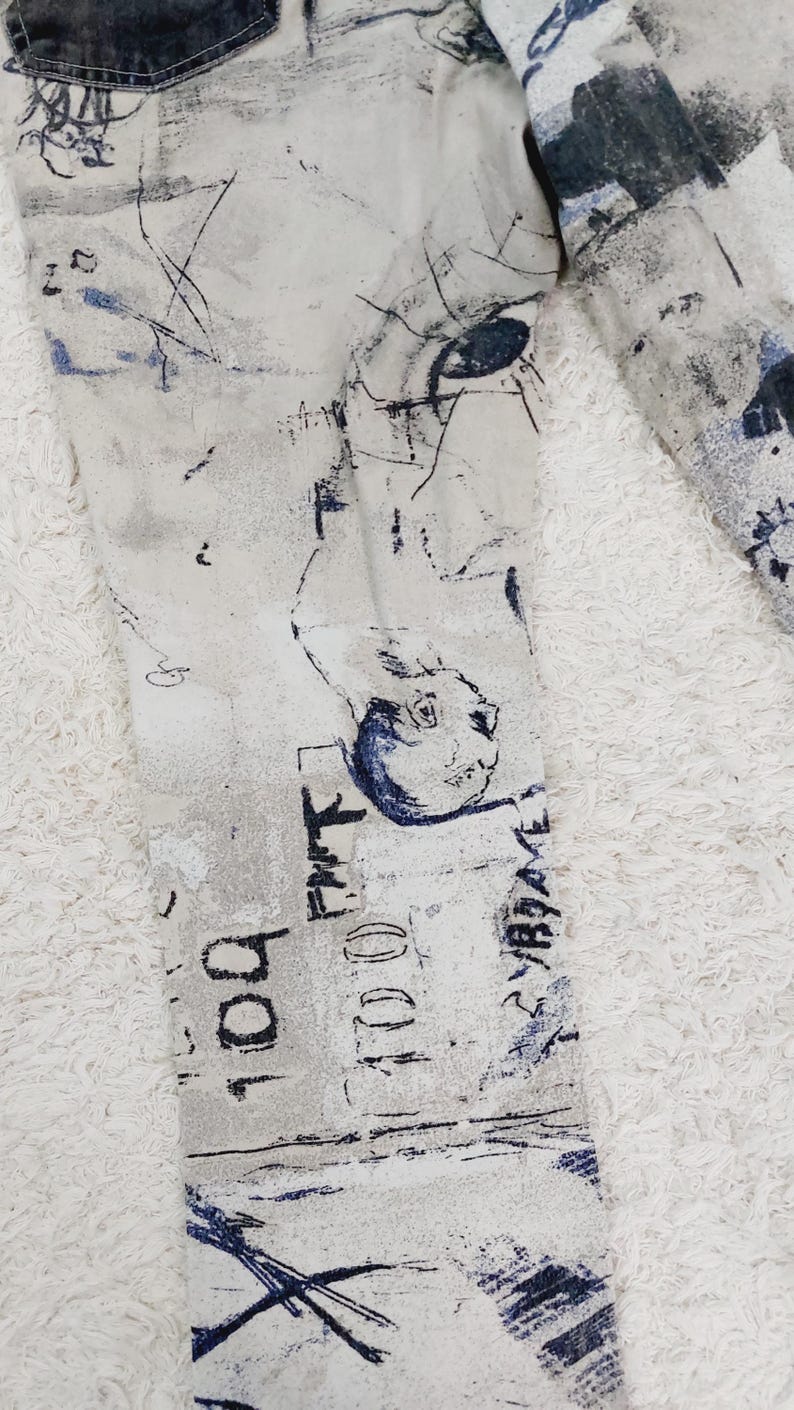 May include: A pair of white denim jeans with a blue and black abstract print. The print features a variety of shapes and lines, including a face with an eye.