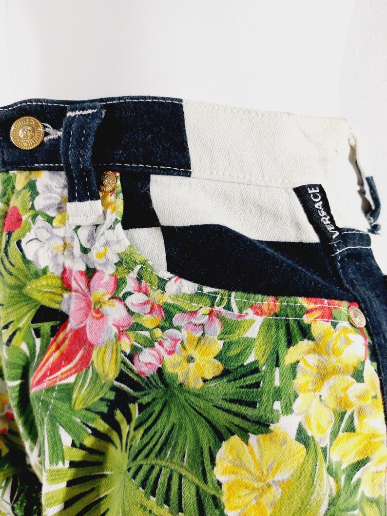 May include: Black and white checkered denim pants with a tropical floral print. The pants have a black waistband with the word "VERSACE" written on the back.