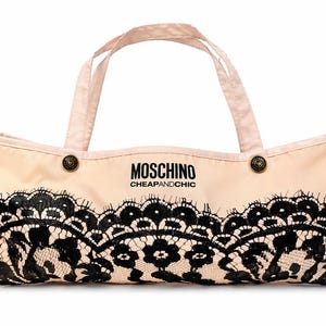 May include: A beige tote bag with black lace detailing along the bottom edge. The bag has two handles and features the text "MOSCHINO CHEAPANDCHIC" in black. Two decorative buttons are on the top of the bag.