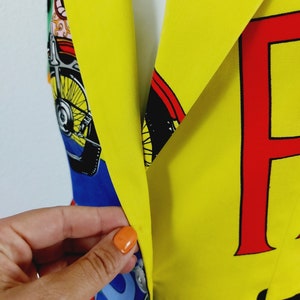 May include: A yellow blazer with a colourful print featuring a motorcycle, a cartoon character, and the letter "F" in red. The blazer has a blue and green patterned lining.