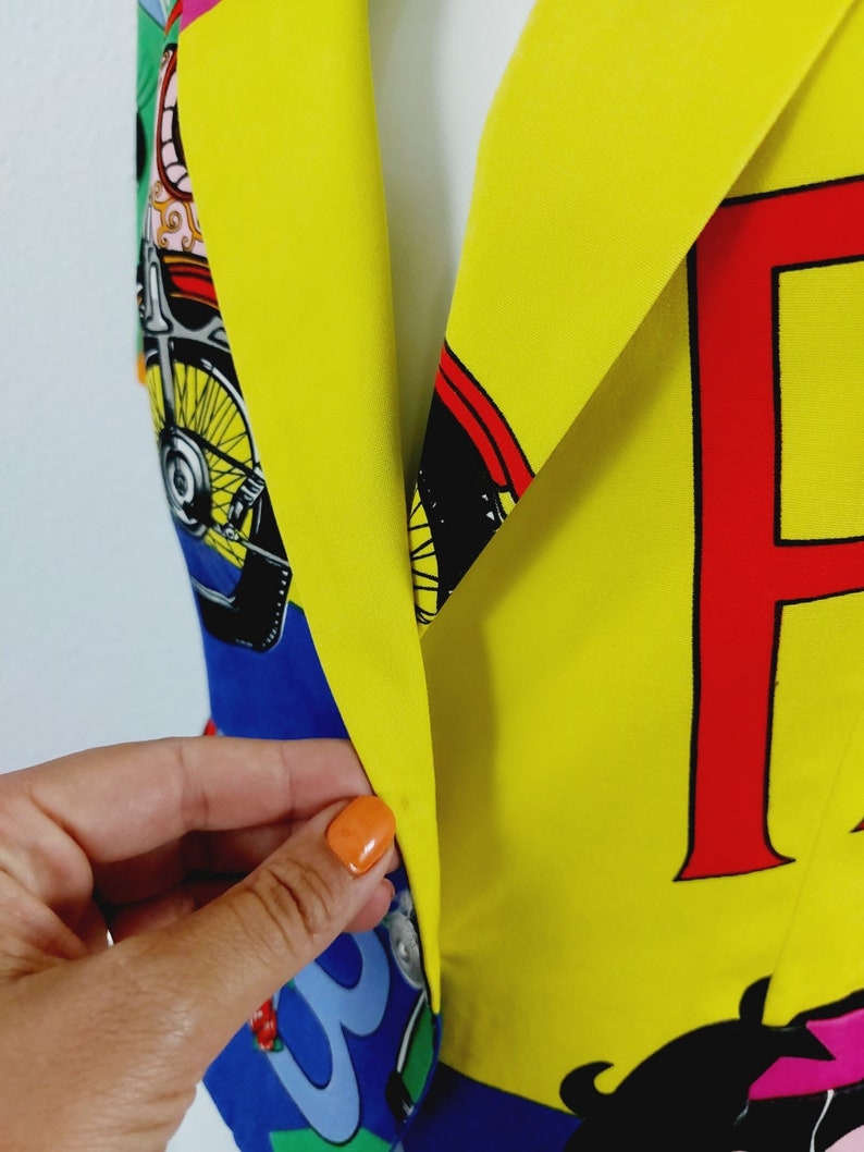 May include: A yellow blazer with a colourful print featuring a motorcycle, a cartoon character, and the letter "F" in red. The blazer has a blue and green patterned lining.