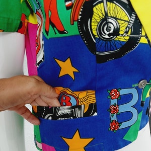 May include: A blue, green, yellow, pink and white patterned short-sleeved button-up shirt with a pocket on the left side. The shirt features a motorcycle, stars and the letter "B" with roses.