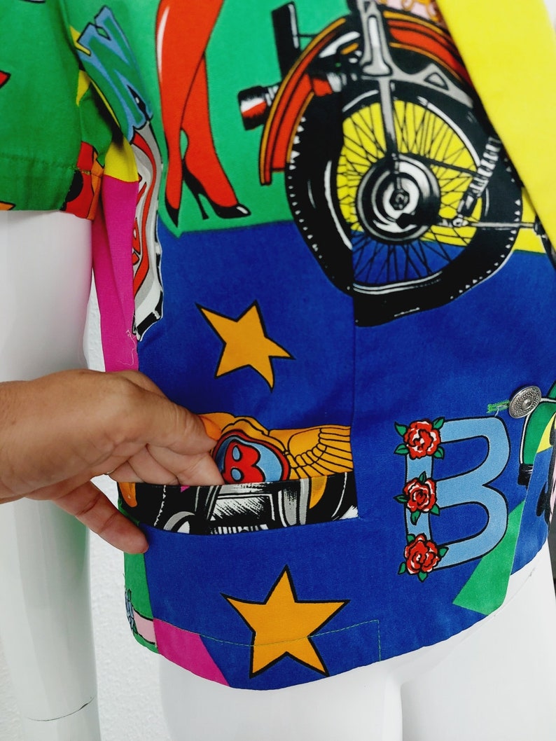 May include: A blue, green, yellow, pink and white patterned short-sleeved button-up shirt with a pocket on the left side. The shirt features a motorcycle, stars and the letter "B" with roses.