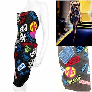 May include: A black strapless denim dress featuring a variety of colorful embroidered patches with the text Versus and VS. The patches are in shades of blue, red, yellow, and white, scattered across the dark fabric in a collage-like arrangement.