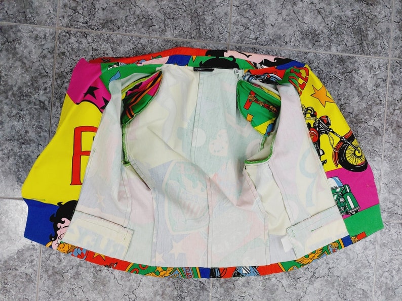 May include: A colourful jacket with a cartoon print featuring a motorcycle, a woman, and the word "FAME". The jacket is unzipped and the lining is white.