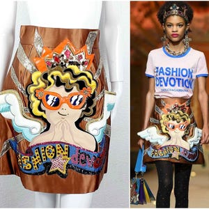 Può includere: Gonna in raso marrone con un design a fumetto di un angelo. L'angelo indossa occhiali da sole arancioni, una corona e ha i capelli ricci gialli. Il testo "FASHION DEVOTION" è visualizzato su uno striscione. La gonna ha un bordo di paillettes.