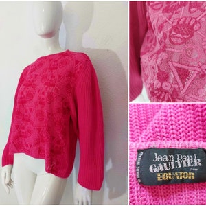 May include: Pink sweater with a textured design and a logo that reads "Jean Paul Gaultier Equator" on the bottom right side. The sweater is made in Italy.